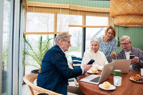 Fototapeta Senior friends using smartphones and laptops at home, cheerful and engaged