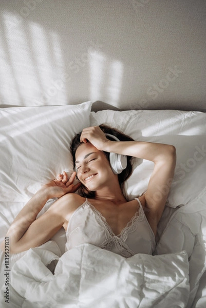 Obraz Woman in headphones lying on bed, listening to music, online streaming and being happy, smile and at home. Young female, girl and with wireless headset being joyful, cheerful or enjoy radio audio