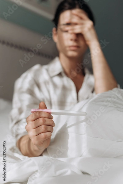 Obraz Young woman seating on bed, holding pregnancy test, displaying a concerned expression, surrounded by soft bedding, evoking feelings of anxiety and hope