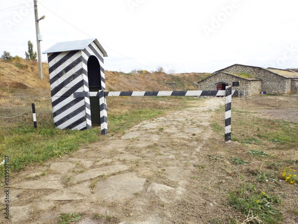 Obraz A guard booth painted in black and white stripes. It looks empty and slanted. In front of the booth is a simple wooden barrier, also painted in black and white stripes. It blocks a narrow stone path.