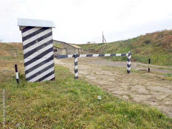 Obraz A guard booth painted in black and white stripes. It looks empty and slanted. In front of the booth is a simple wooden barrier, also painted in black and white stripes. It blocks a narrow stone path.