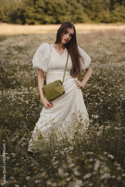 Obraz Charming Woman in Long Boho Lace Dress with Small Green Crossbody Handbag Standing Among Blooming Chamomile Meadow