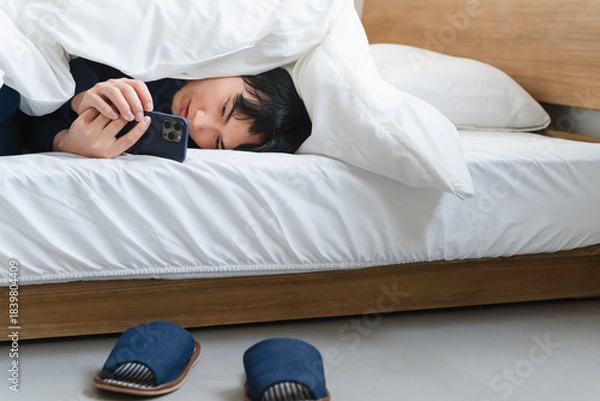 Fototapeta 布団にくるまりスマートフォンを操作している若い男性　ライフスタイルイメージ