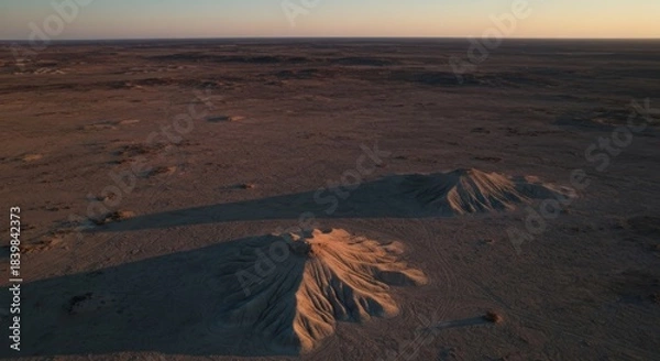 Fototapeta Aerial view of two eroded, sand-like mounds in a vast, desolate landscape at sunset