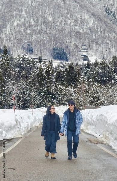 Obraz ​A couple walks hand-in-hand down a snow-covered mountain road, flanked by deep snowdrifts and a dense, snowy forest backdrop in winter