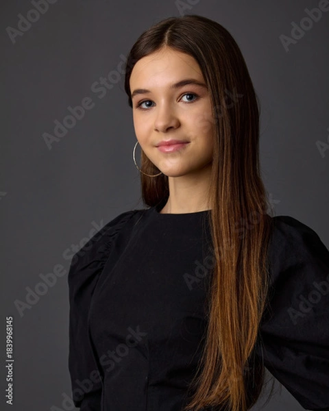 Fototapeta A close-up studio fashion portrait of a young woman wearing a black top with voluminous sleeves and large hoop earrings. Against a dark gray background, the focus is on her beauty and hairstyle.