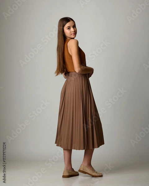 Fototapeta A studio fashion portrait of a young woman, seen from behind and in profile, wearing a monochrome brown ensemble consisting of a pleated midi skirt, a simple blouse, and loafers. Posing against a ligh