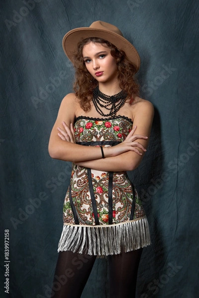 Fototapeta Close-up of a fashionable portrait of a young woman in a strapless dress with a bright floral print in a folk style. She is wearing a brown fedora hat and a black beaded necklace.