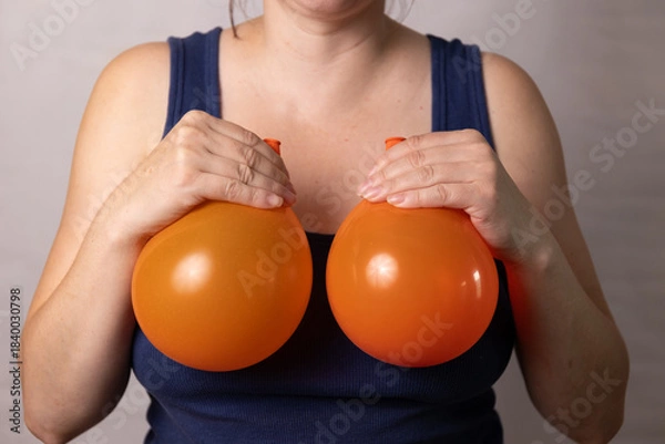 Obraz A woman holds two orange balloons in front of her chest, representing the effects of pregnancy and age on breasts. She examines the changes while reflecting on options for body image improvement.