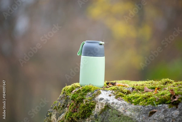Fototapeta Thermos flask with hot coffee or tea drink is standing on a moss covered rock in a forest, traveling and wanderlust concept, food container