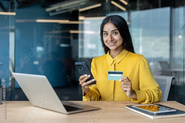 Fototapeta Portrait of a smiling young Muslim woman sitting at a desk in the office, using a phone and credit card, looking at the camera