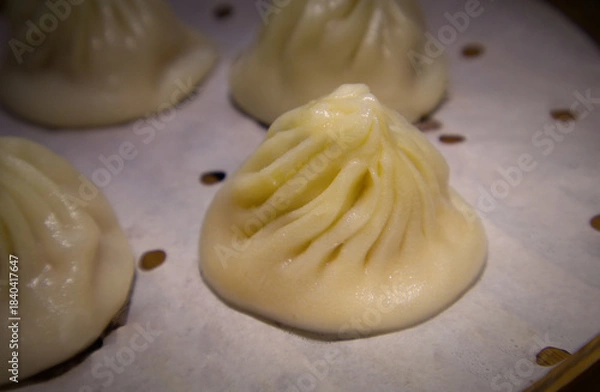 Fototapeta A close-up of a single Xiao Long Bao (soup dumpling) with its characteristic pleated top, sitting on a parchment liner in a steamer basket. 