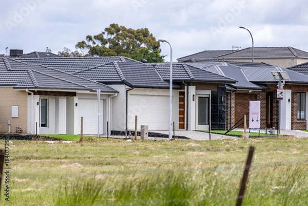 Obraz New single-storey homes in an outer-suburban Melbourne estate bordered by vacant grassland. Concept of urban expansion, residential development, suburban housing market, and undeveloped land buffer
