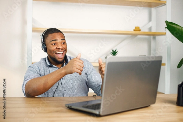 Fototapeta Black man sitting in office, wearing a earphones and make a video call