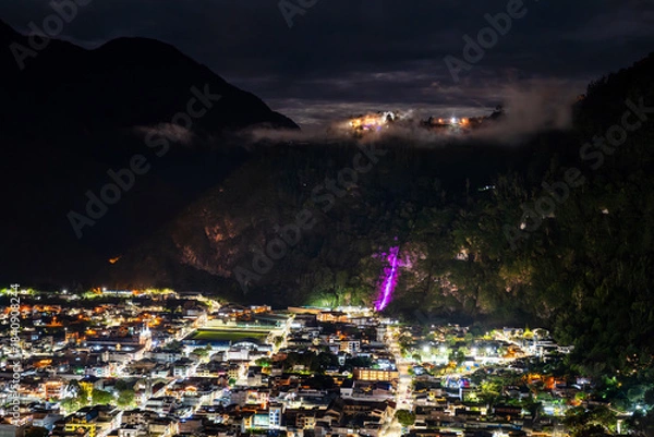 Fototapeta Banos de Agua Santa town center glows at night in Ecuador. Panoramic view features illuminated Cascada de la Virgen waterfall on cliffside above city streets