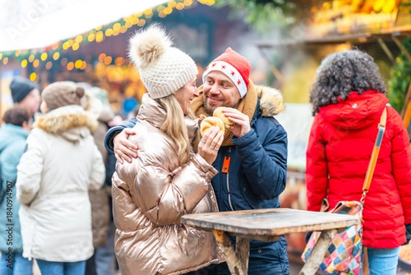 Fototapeta Smiling couple eating warm sausages and rolls at a Christmas market surrounded by glowing festive lights