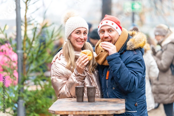 Fototapeta Smiling couple sharing sausages and rolls at a Christmas market enjoying festive winter food and bright lights
