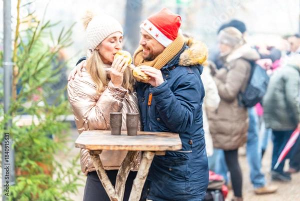 Fototapeta Cheerful couple enjoying warm sausages and rolls at a Christmas market filled with winter charm and lights
