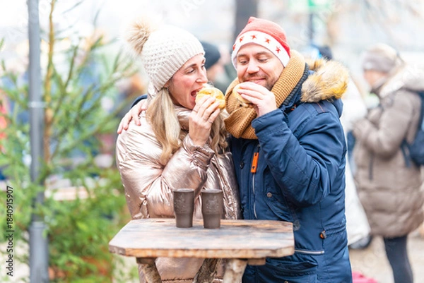 Fototapeta Happy couple enjoying tasty sausages and rolls at a Christmas market surrounded by warm seasonal lights