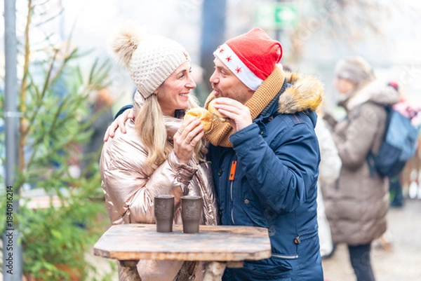 Fototapeta Smiling couple enjoying sausages and rolls at a Christmas market filled with winter warmth and festive charm