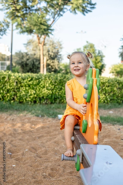 Fototapeta young girl with blonde hair kghappily sits on playground seesaw