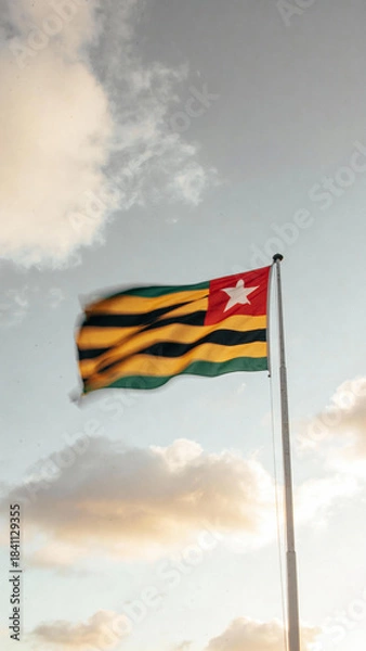 Obraz A flag with horizontal stripes of green and yellow, featuring a red square with a white star, waving against a backdrop of clouds and a soft sky.