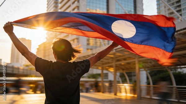 Obraz Silhouette of a person proudly holding the Laotian flag against a colorful sunset, ideal for celebrating national pride in media or social posts.