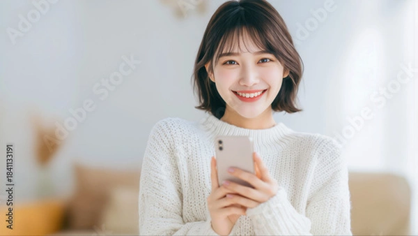 Obraz 明るいリビングで白いニットを着てスマートフォンを持ち笑顔を見せる若いアジア人女性