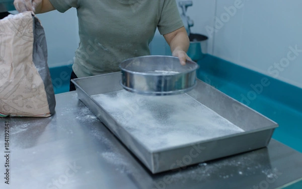 Fototapeta Unknown hand people sifting flour with sifter on stainless steel table in clean food preparation area, Factory production line baking preparation professional kitchen workflow industrial technology.