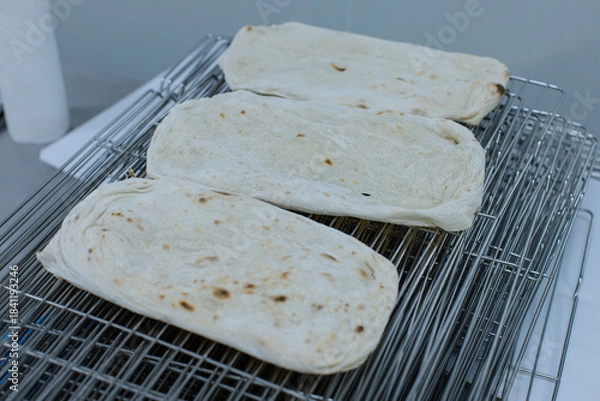 Fototapeta Freshly baked pizza rectangular flatbreads rest on stainless steel cooling rack light golden spots soft texture clean, professional baking workflow environment production process technology factory.