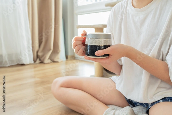 Obraz Minimalist morning moment at home: a young person sitting on the floor and holding a ceramic mug with both hands. Soft natural light, cozy atmosphere, relaxation and mindfulness concept.