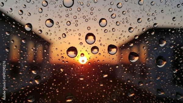 Fototapeta Mesmerizing close-up of raindrops clinging to a window pane, perfectly framing a blurred, warm sunset between residential buildings, creating a serene and reflective atmosphere