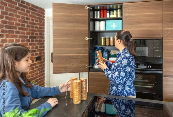 Obraz Mother and daughter adding cookies packages to emergency pantry closet including water, canned food and first aid kit. Family preparing 72 hour survival kit for natural disaster or power outage