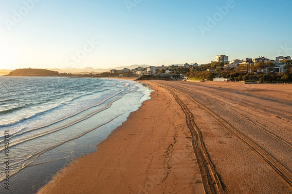 Fototapeta Drone view of wide sandy beach with gentle waves, morning sunlight, seaside and distant city skyline