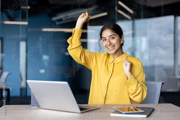 Fototapeta Portrait of a young Indian woman sitting at a desk in the office, rejoicing and dancing, looking at the camera with a smile