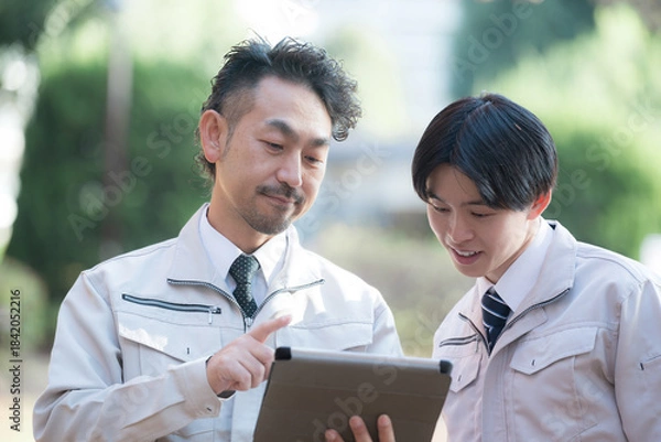 Obraz 新緑の屋外の施設管理など現場で働く作業着を着た施工管理士などのビジネスマン　タブレットを見る