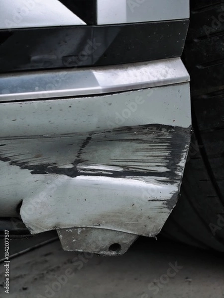 Obraz A vertical close-up of a severe accident, impact, or scrape damage to the silver plastic front bumper fender of a car, showing deep black scratches, cracks, and distortion near the wheel