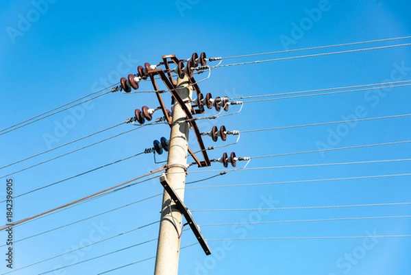 Obraz Old concrete utility pole with multiple electric power lines and insulators, set against a vibrant clear blue sky. Ideal for energy, infrastructure, and communication concepts.