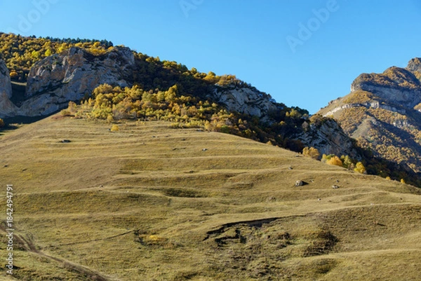 Obraz Beautiful landscape featuring rolling hills and towering rock formations under clear blue sky, with golden autumn foliage adding vibrant contrast. Kabardino-Balkaria. El-Tyubu