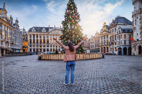 Fototapeta A happy tourist woman stands in front of a beautiful decorated Christmas Tree for the festive season at the Grand Place in Brussels, Belgium