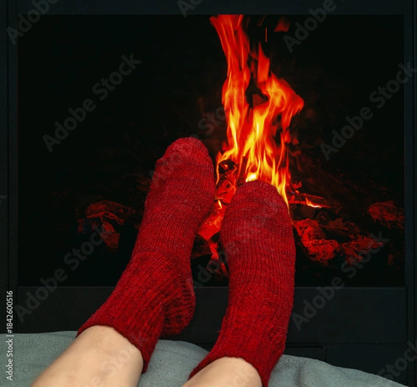 Fototapeta Winter scene with legs in red woolen socks resting near glowing fireplace. Feelings of relaxation.