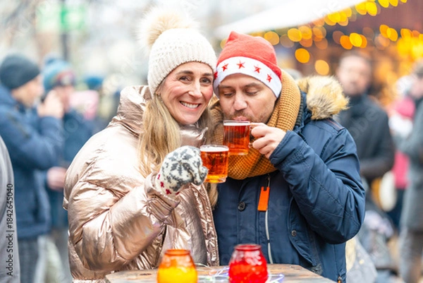Fototapeta Smiling couple with mulled wine and punch mugs standing before a Christmas market glowing with holiday lights