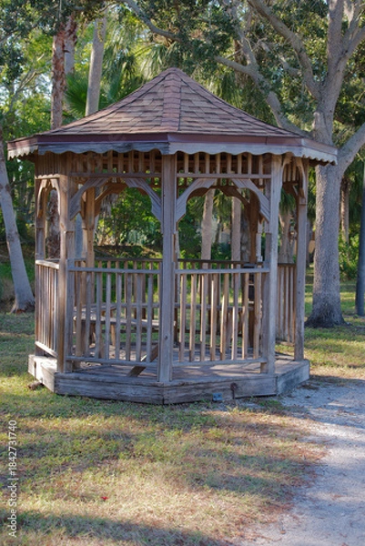 Obraz Wooden Gazebo and Park Bench in a Lush Urban Park Setting. Rustic wooden gazebo and a vibrant orange park bench under a lamppost in a green park. The peaceful ambiance and bright vegetation symbolize 