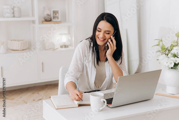 Obraz A woman sits at a desk with laptop talking on phone, smiling