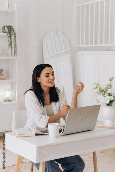 Obraz Top view of brunette woman typing text on a laptop on white office desk with table lamp, notebook, bottle of drinking whater and flowers in vase. Concept of freelancing, remote work, online learning