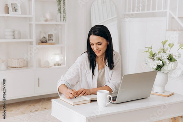 Obraz Young brunette freelancer woman sitting at desk using laptop writing notes while studying online, watching webinar, looking at pc screen learning web classes or remote working from home. Side view