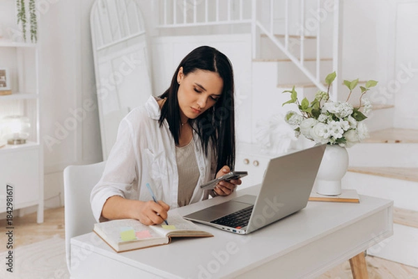 Obraz Independent woman at workstation, Female freelancer organizing her daily schedule and goals at desk, Independent woman multitasking with technology and planning tools in serene home office setting