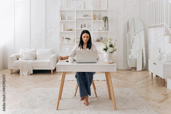 Obraz Caucasian freelancer woman typing and referencing notes, creative copywriter editing drafts beside phone and notebook, white desk with floral accents and bright natural light, thoughtful mood