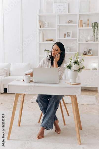 Obraz Caucasian freelancer woman on phone, remote project manager taking client notes at bright white desk with laptop, mug, minimalist bookshelf and natural light, focused professional mood