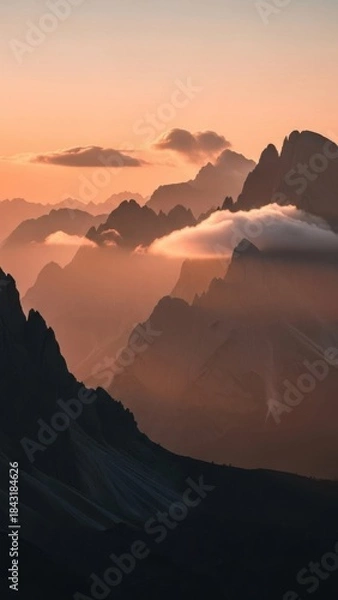 Obraz Layers of majestic mountains at sunrise, with soft clouds nestled between peaks under a warm, gradient sky.